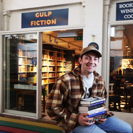 Picture of the front of Gulp Fiction shop with owner in the foreground holding books