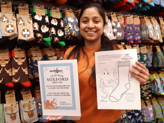 Female with a big smile standing infront of a wall of socks. She is holding up the entry sheet for the design a sock competition