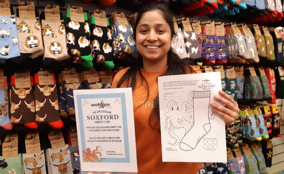 Person holding up sock design entry forms and smiling to camera. Standing in front of a wall of socks