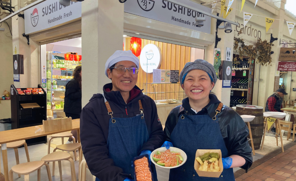 Two people smiling at us, standing in front of sushi bowl business premises and holding up sushi dishes