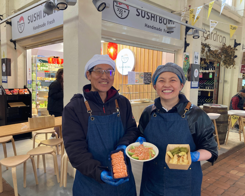 Two people smiling and standing in front of Sushi Bowl shop. Each person is holding up a sushi dish