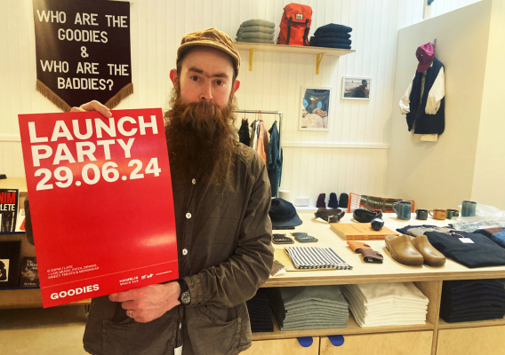 Matt owner of Goodies holds poster graphic with event details, in his shop surrounded by mens clothing